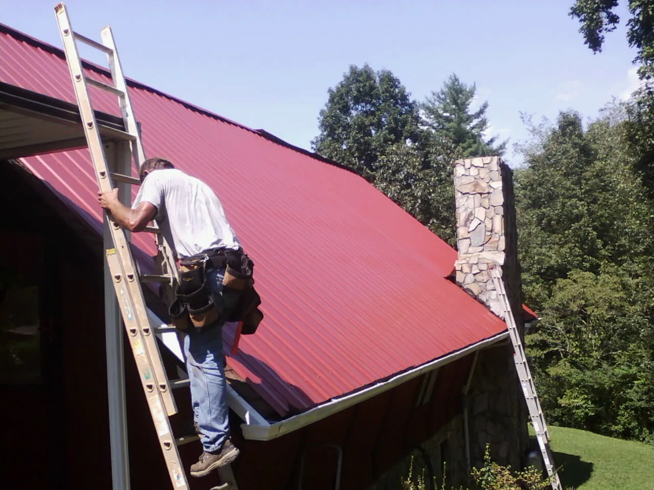 Roofer on a ladder beside a red metal roof and stone chimney — Soffit & Fascia Repair by Roofing Brothers in Villa Park